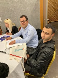 Yuvarlak bir masanın etrafında oturan üç kişi var. Soldaki kadın sarı saçlı ve siyah bir ceket giymiş, önünde bir defter var ve yazı yazıyor gibi görünüyor. Ortadaki kişi Oğuz ÖZDEMİR gözlüklü ve mavi bir gömlek giymiş, önünde bazı belgeler var. Sağdaki genç kişi Mert YAKIŞTIR siyah bir deri ceket giymiş ve elinde beyaz bir baston tutuyor. Masanın üzerinde "Vestel" yazılı bir bez çanta var. Arka planda gri bir duvar ve açık bir kapı görülüyor.